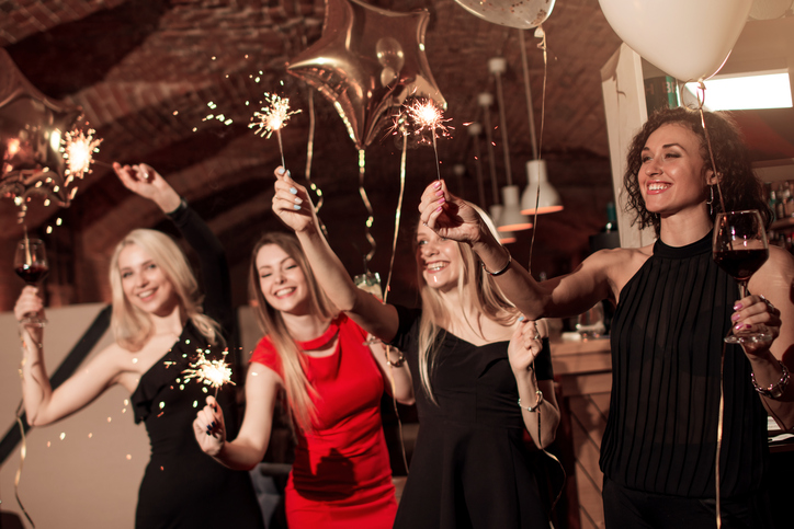 Group of happy smiling girlfriends wearing evening dresses celebrating New Year holding sparklers in decorated cafe