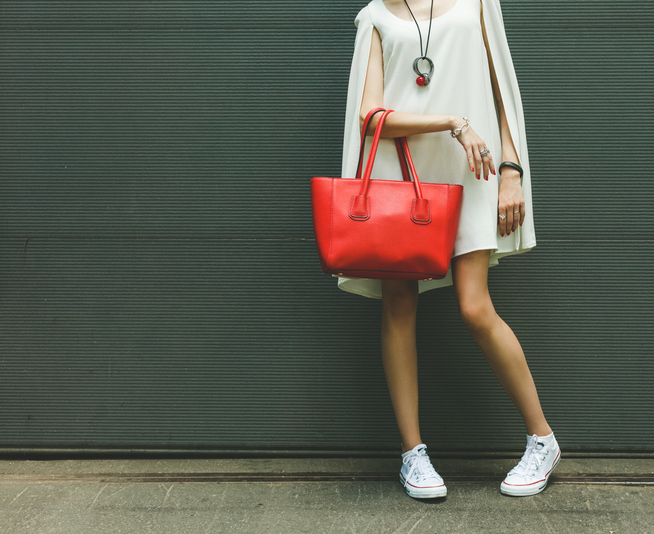 Fashionable beautiful big red handbag on the arm of the girl in a fashionable white dress and sneakers, posing near the wall on a warm summer night. Warm color. big red handbag on the arm of the girl
