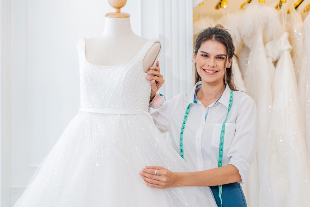 Smiling caucasian woman is bridal shop owner tidying up the wedding dress and looking at camera at wedding studio, Small business entrepreneur wedding planner and tailor designer concept